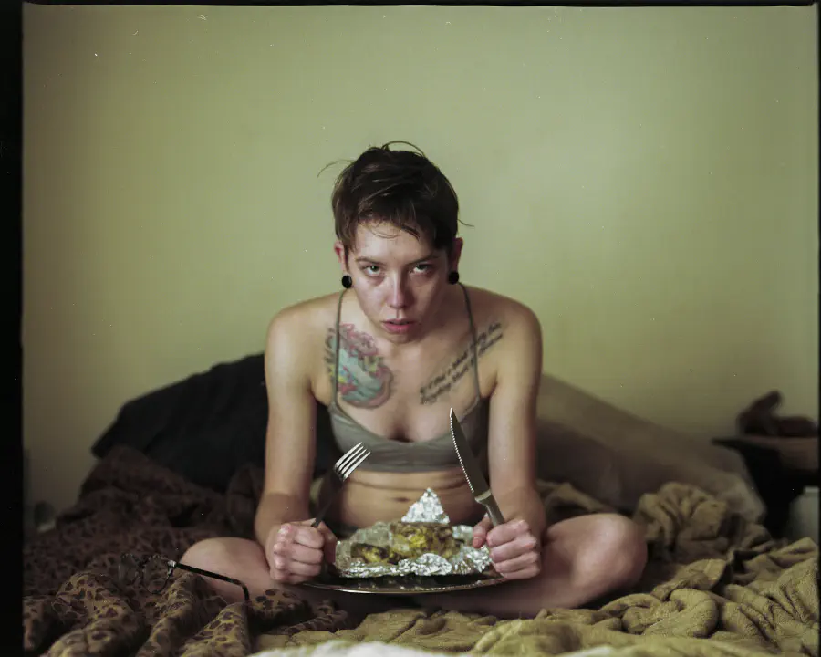 A woman sitting inher underwear holding a tray with rotten food. She is holding a fork and knife and stares into the camera with her mouth slightly ajar.