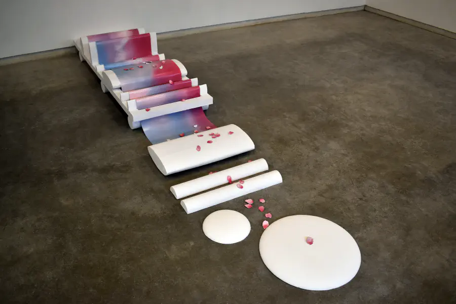 A white platform has matte paper with pink and blue gradients folded and wrapped on top of it. Foam cylindrical 'stepping stones' lead to 2 white oval 'stones.' Pink flower petals are scattered across the structure, seemingly a path.