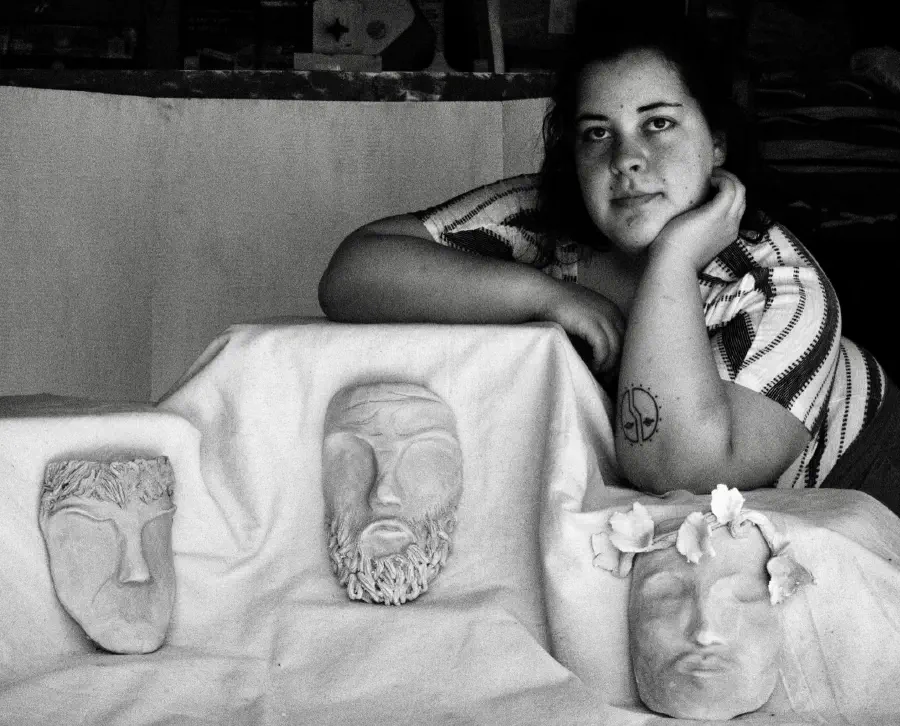 A person with her arms propped up as she sits behind several expressive clay sculpture masks. None have eyes, but they all have eyebrow ridges to suggest expressions.