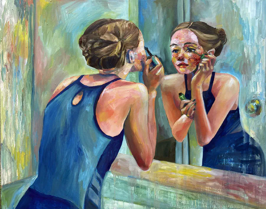 A girl with her hair up wearing a blue dress peers into the mirror as she puts on mascara.