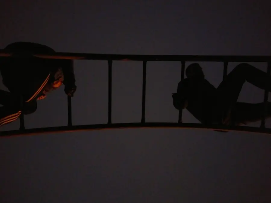 A photo from the ground-up depicting two people on a structure, perhaps monkey bars. One person is crawling towards the other.