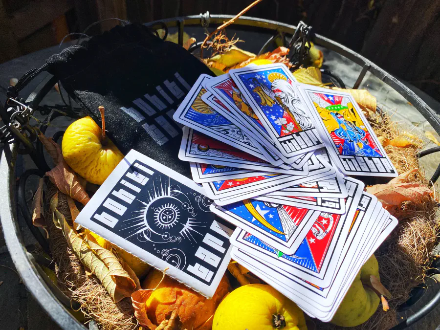 A stack of tarot cards on a pile of yellow and orange squash and hay. The cards depict Puerto Rican morifs and figures.