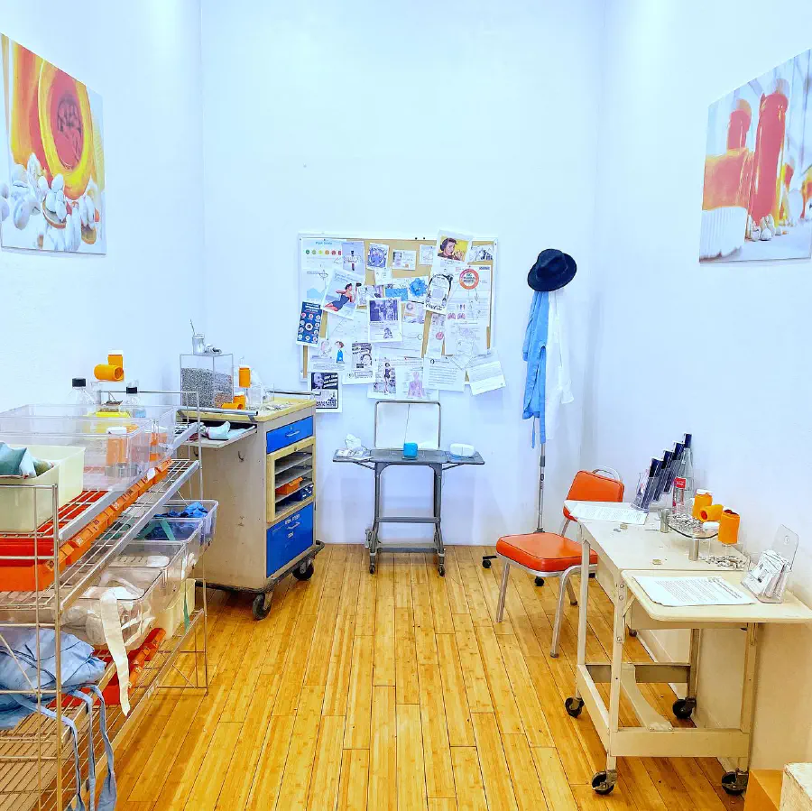 A bright white room that seems to be an art studio. A corkboard at the end of the room has a variety of papers pinned to it. The room has a lot of orange and blue.