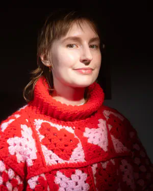 An image of a person with blond hair and a thick-knit pink, white, and red heart-themed sweater smiling gently.